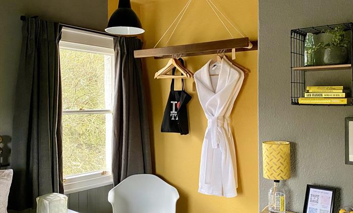 A yellow and grey bedroom, with a chair in the corner, chest of drawers and yellow lamp, and a bathrobe hanging on a yellow wall, at Crafnant House, B